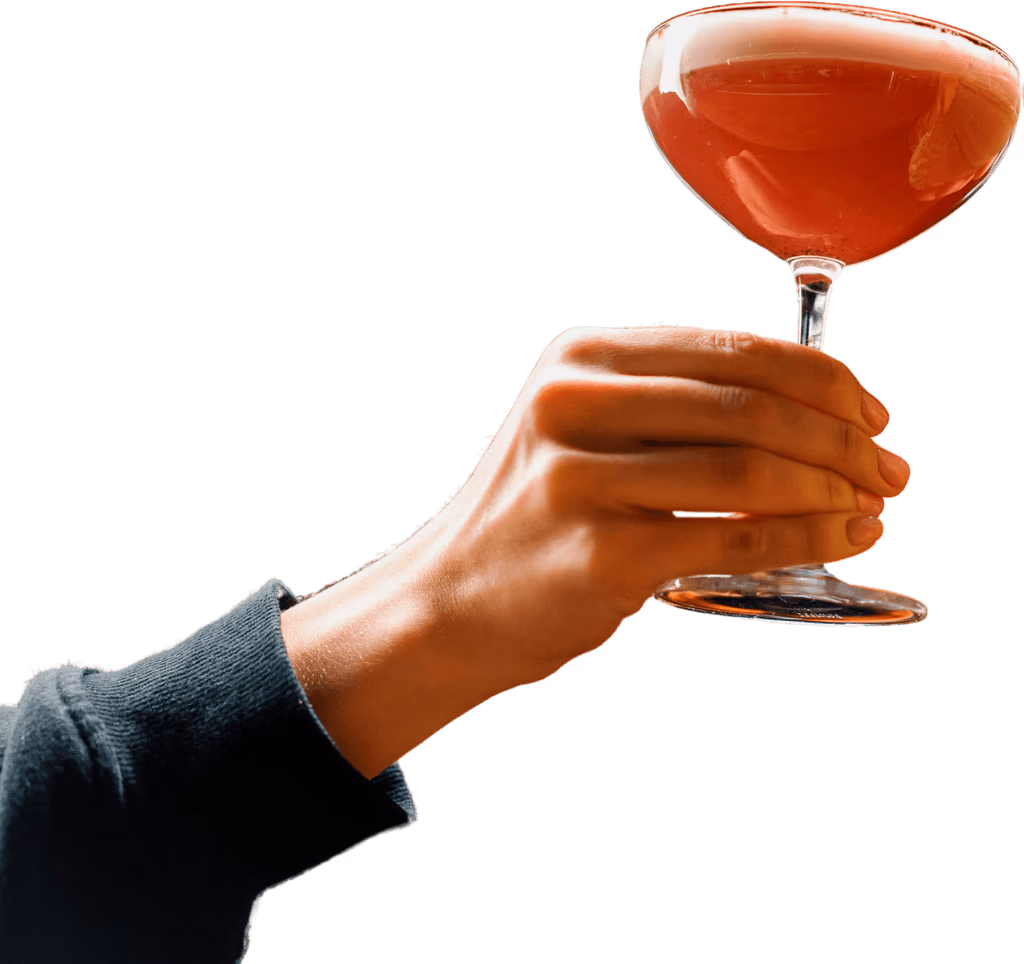 A hand holding a cocktail glass filled with a reddish drink against a neutral background.